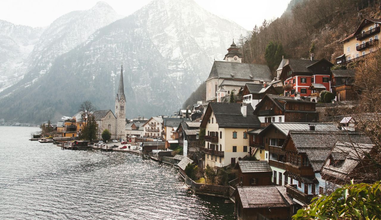 A lakeside village features traditional alpine houses, a tall-steepled church, and snow-covered mountains in the background.