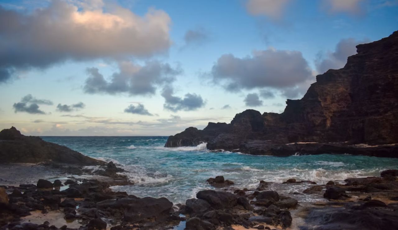 Rocky coastline with crashing waves and dramatic cliffs under a colorful evening sky with scattered clouds.