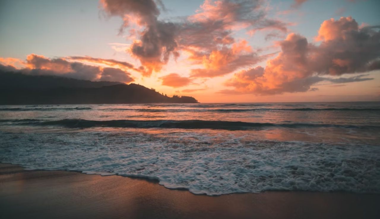 Vibrant sunset over Hanalei Bay in Hawaii, with waves gently washing onto the shore and dramatic clouds lighting up the sky.