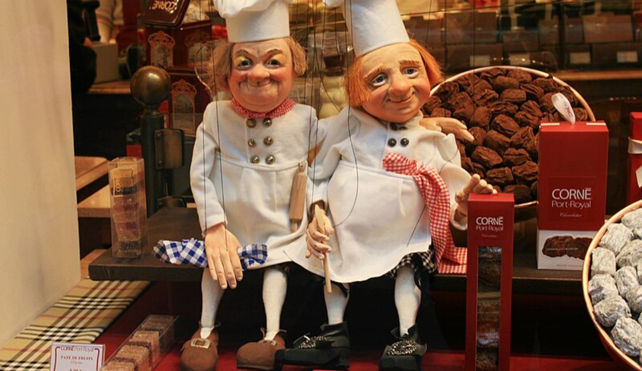 Window display of a Belgian chocolate shop featuring two cheerful chef dolls surrounded by boxes of chocolate truffles and sweets.