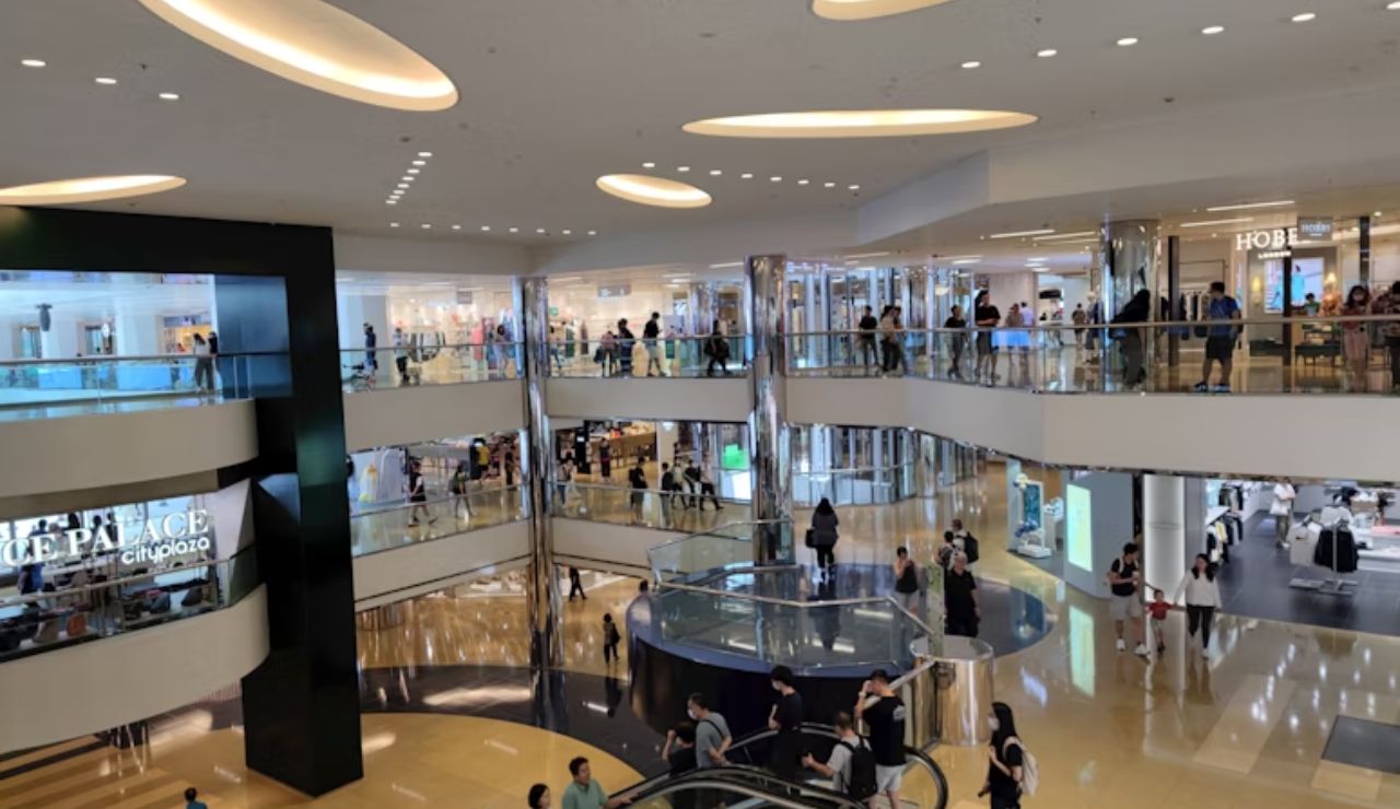 Modern multi-level shopping mall interior with escalators, glass railings, and shoppers walking through various stores.