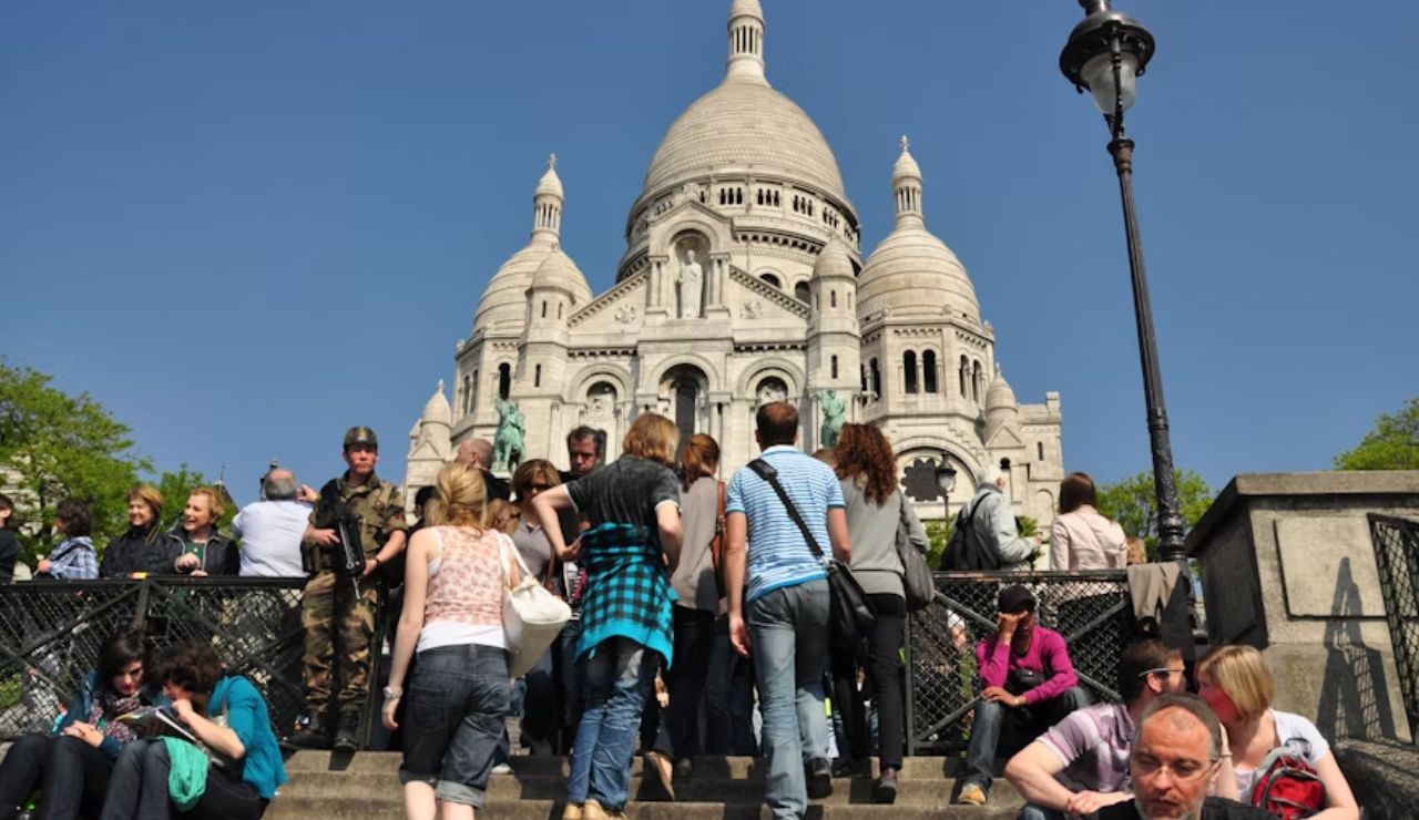 Tourists gathered on the steps leading to the Sacré-Cœur Basilica in Paris, with the white-domed church prominently visible under a clear blue sky.