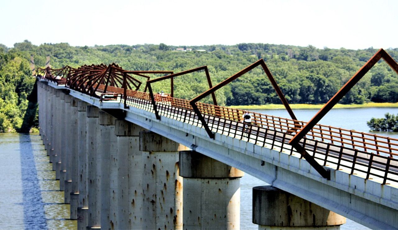 Cyclist rides across a long elevated bike bridge with angular metal supports over a wide river.