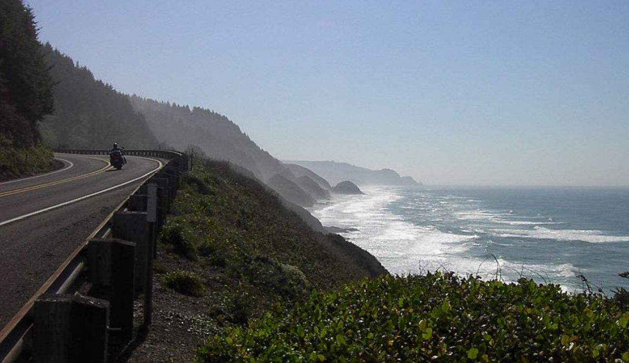 Motorcyclist riding along a winding coastal highway overlooking the Pacific Ocean, with rugged cliffs and sea mist in the distance.