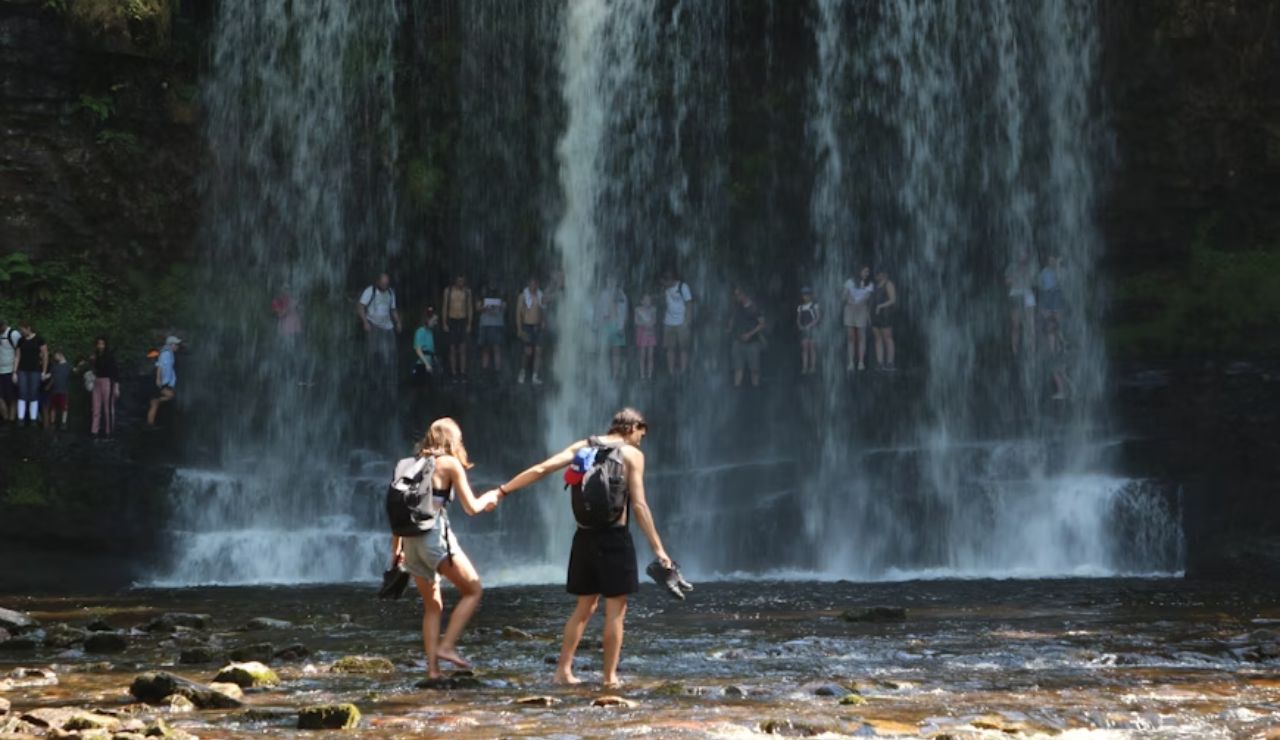 Couple holding hands while walking barefoot near a waterfall with onlookers behind.
