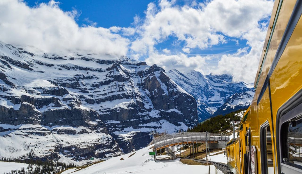 A yellow train traveling through a snowy mountainous landscape under a blue sky with clouds.