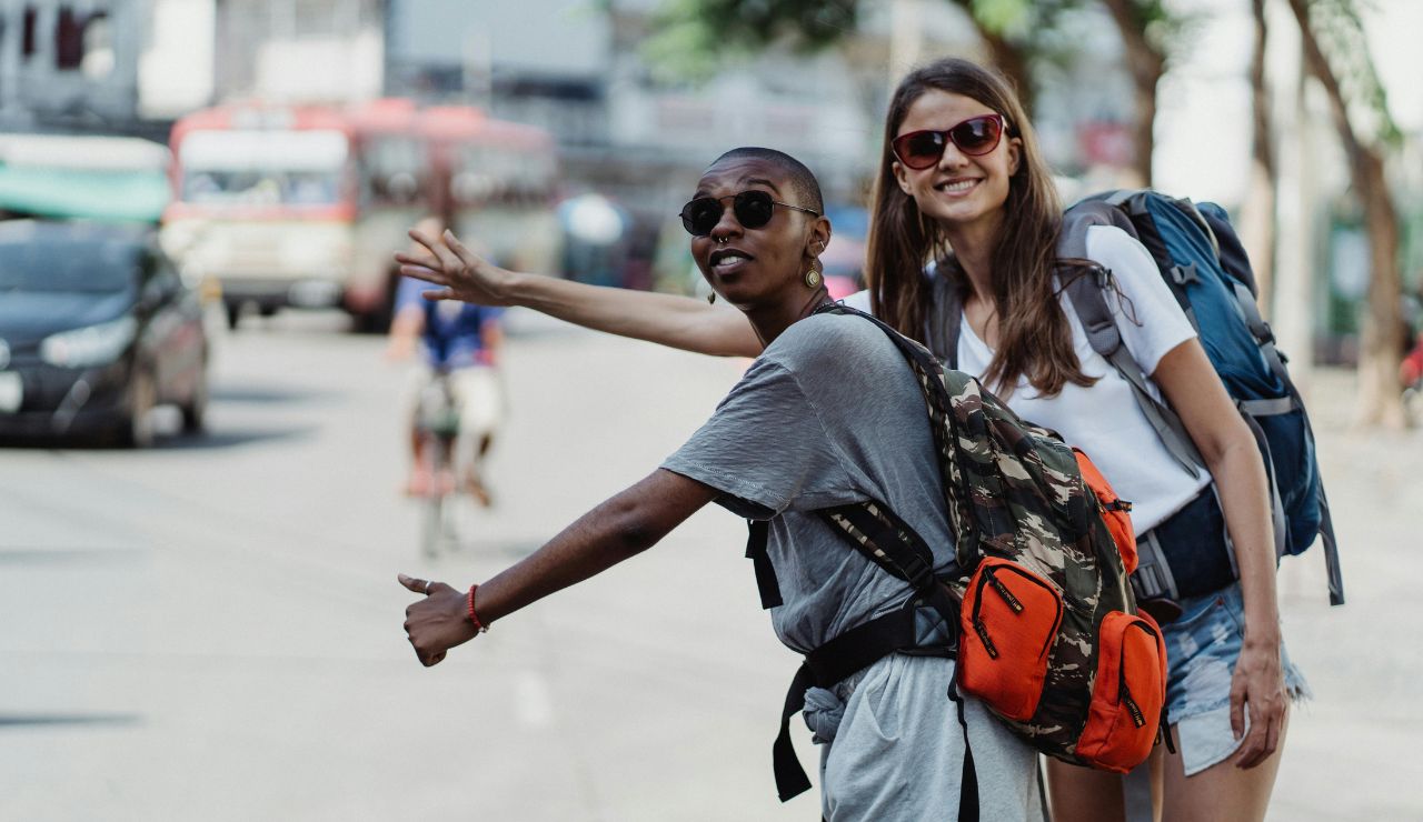 Two smiling backpackers hitchhiking on a city street, holding out their thumbs and wearing sunglasses.
