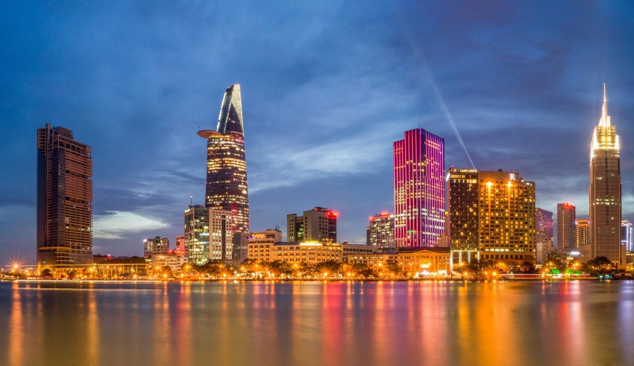 Night view of a modern city skyline with illuminated skyscrapers reflecting on a calm river.