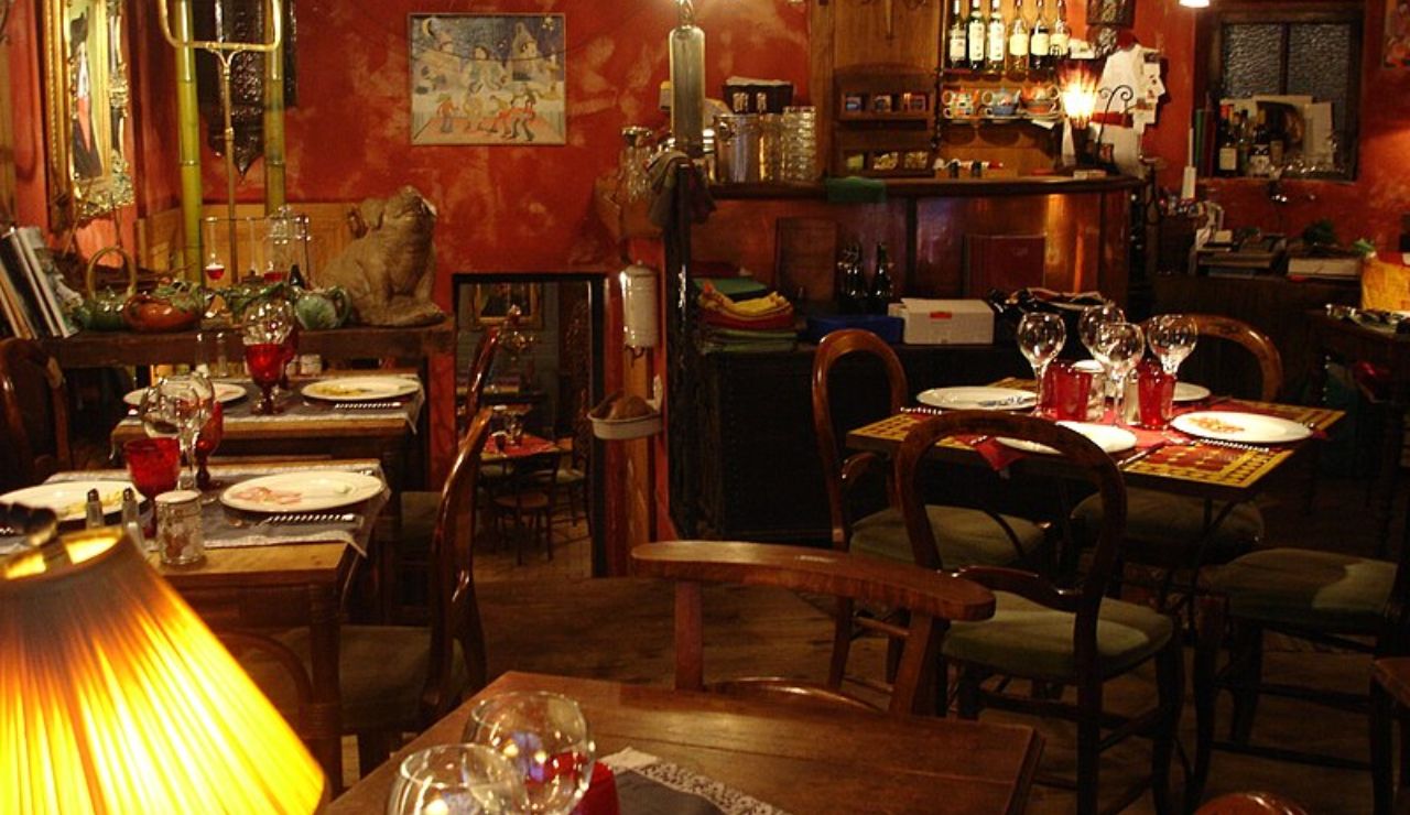 A cozy, dimly lit restaurant interior with rustic wooden tables, red walls, and vintage decor. Tables are set with wine glasses, plates, and red-tinted glassware.