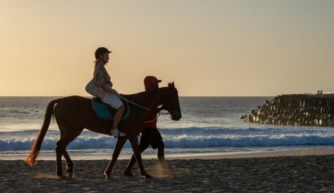 Person riding a horse along the beach at sunset with a guide.