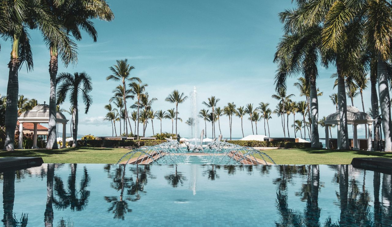 A large pool reflects palm trees and a metal dolphin sculpture beneath a blue sky, flanked by grassy areas and gazebos.