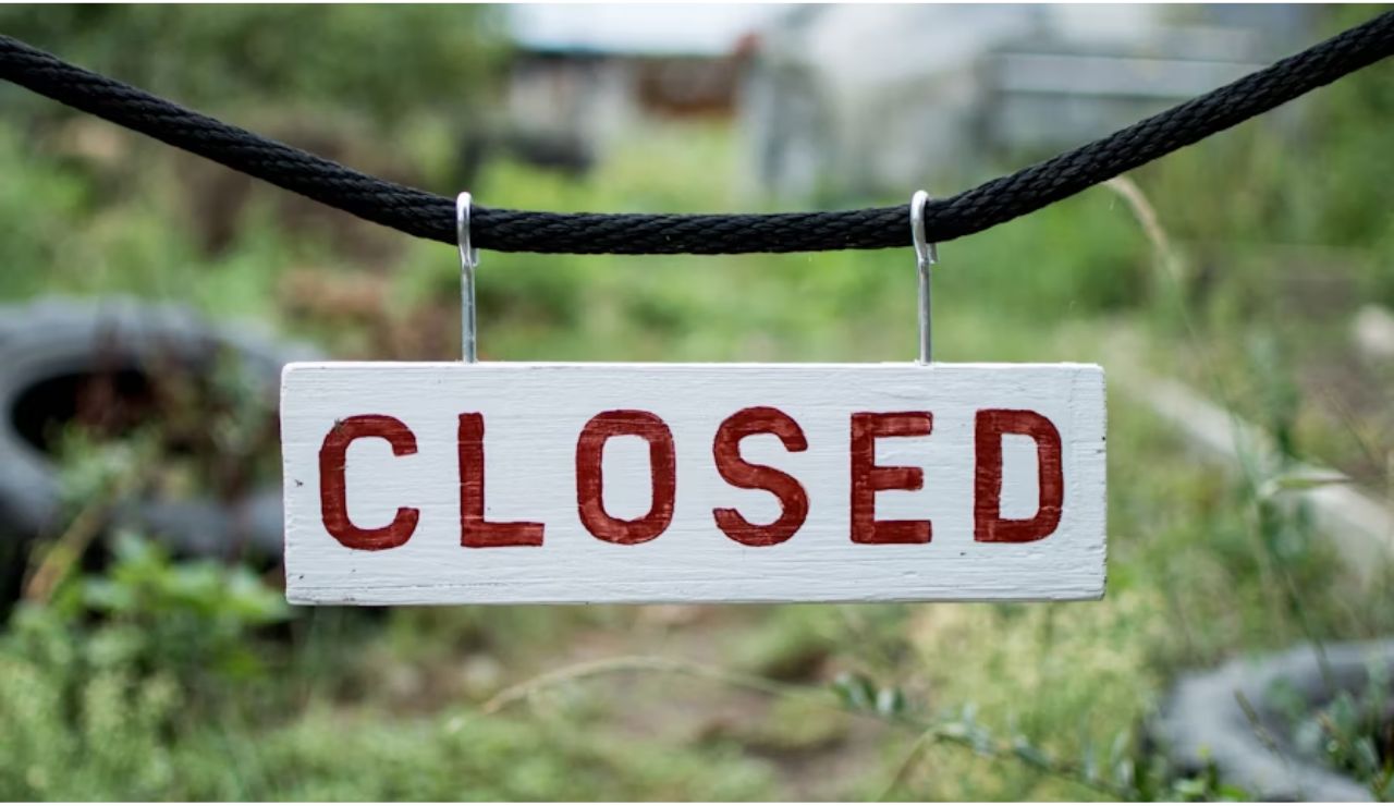 White wooden sign with red letters reading "CLOSED" hanging on a black rope in an overgrown outdoor area.