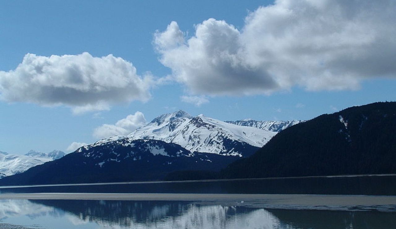 Snow-capped mountains reflect on the calm waters of Turnagain Arm in Alaska under a bright blue sky with scattered clouds.