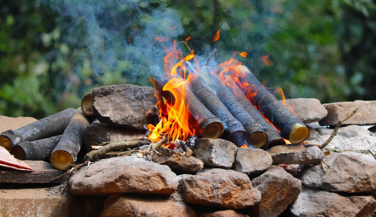 Close-up of bamboo logs burning over a stone fire pit with flames and smoke rising in a forest setting.