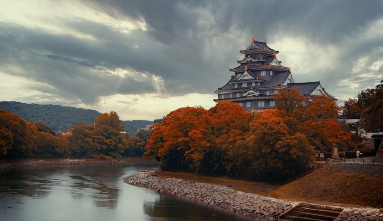 Traditional Japanese castle surrounded by vibrant autumn trees beside a calm river under a dramatic sky.