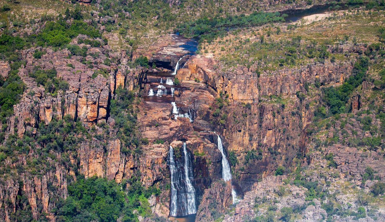 Tall, multi-tiered waterfall cascading down rugged, rocky cliffs surrounded by sparse vegetation and greenery in a remote landscape.