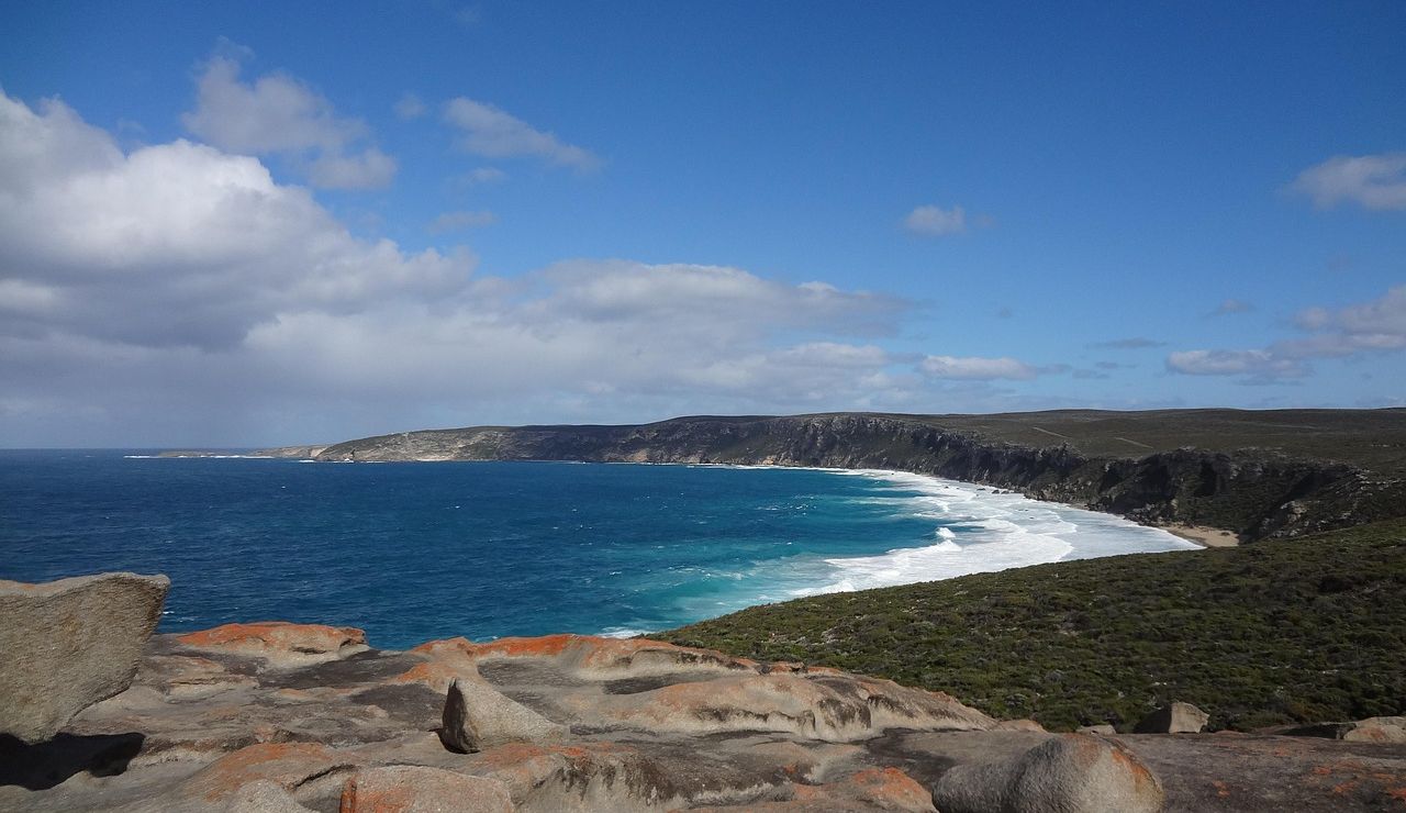 Rocky coastal viewpoint overlooking a sweeping bay with rugged cliffs, turquoise ocean waves, and a partly cloudy sky.