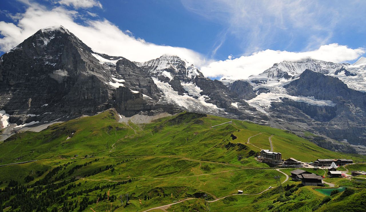 Lush green alpine meadows stretch beneath towering snow-capped mountains, with scattered buildings and winding trails under a bright blue sky.