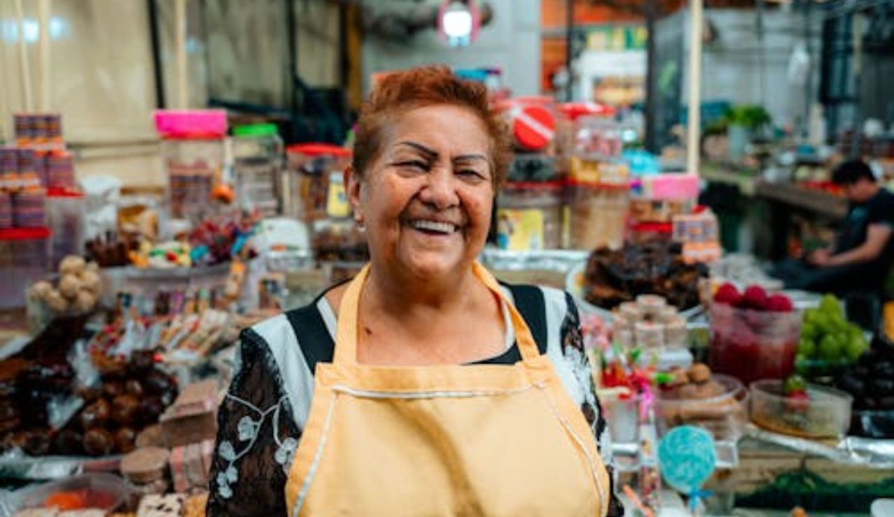 Smiling woman wearing an apron standing in front of a colorful market stall filled with sweets and snacks.