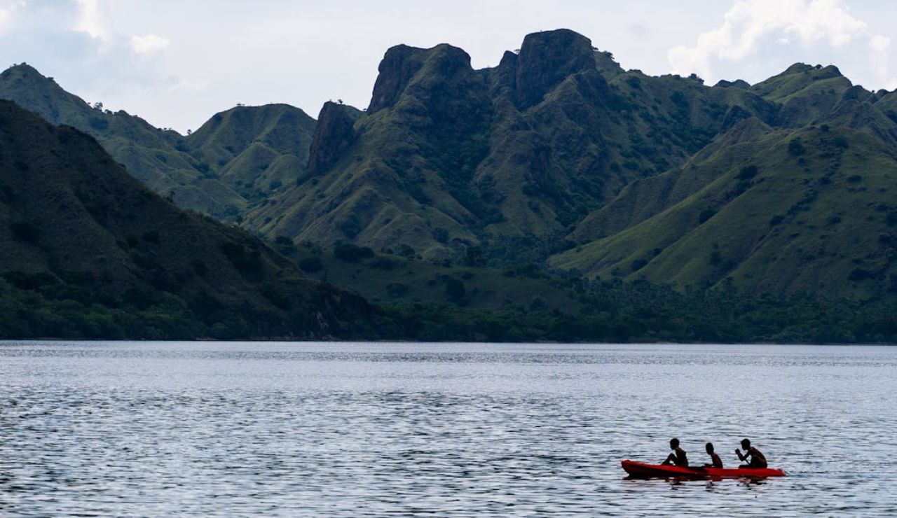 Three people paddle a red kayak across a calm lake with lush, rugged mountains in the background.
