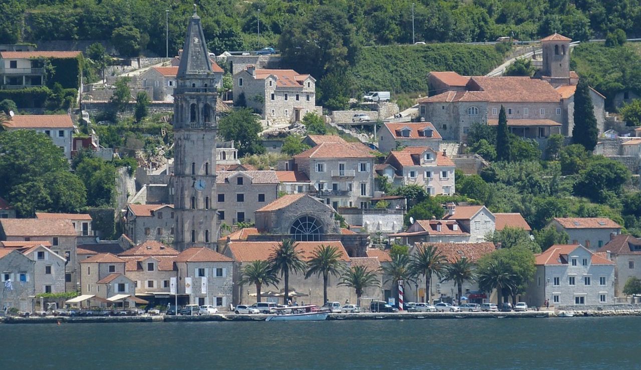 Coastal town with stone buildings and red-tiled roofs, dominated by a tall bell tower and lined with palm trees along the waterfront.