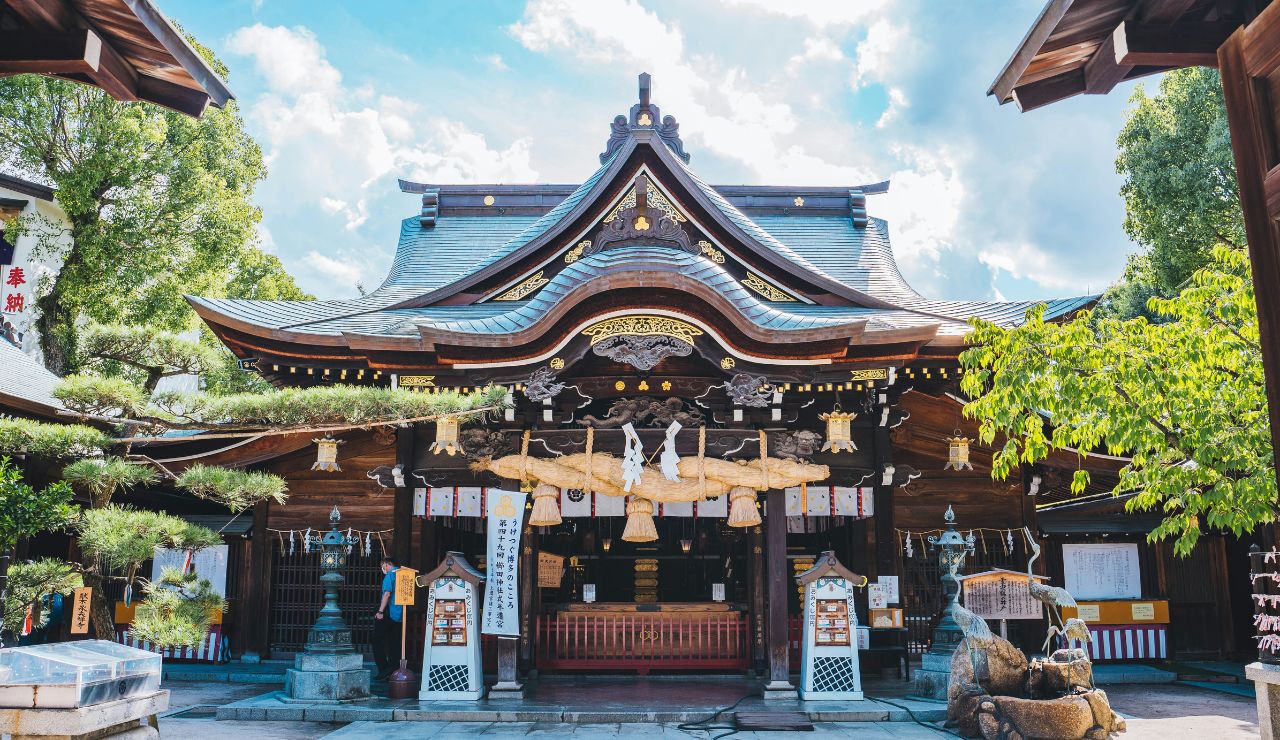 Traditional Japanese Shinto shrine with ornate wooden architecture, decorative roof, hanging ornaments, and trees beneath a partly cloudy sky.