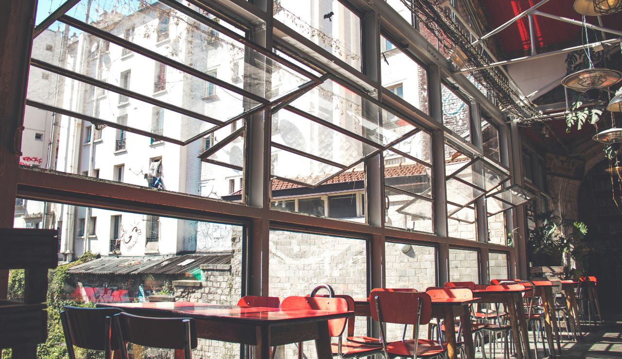 A bright café with empty red chairs and wooden tables sits by large open windows overlooking city buildings.