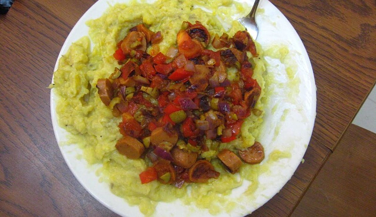 Plate of mashed green plantains (mofongo) topped with a colorful mix of sautéed vegetables and sausage.