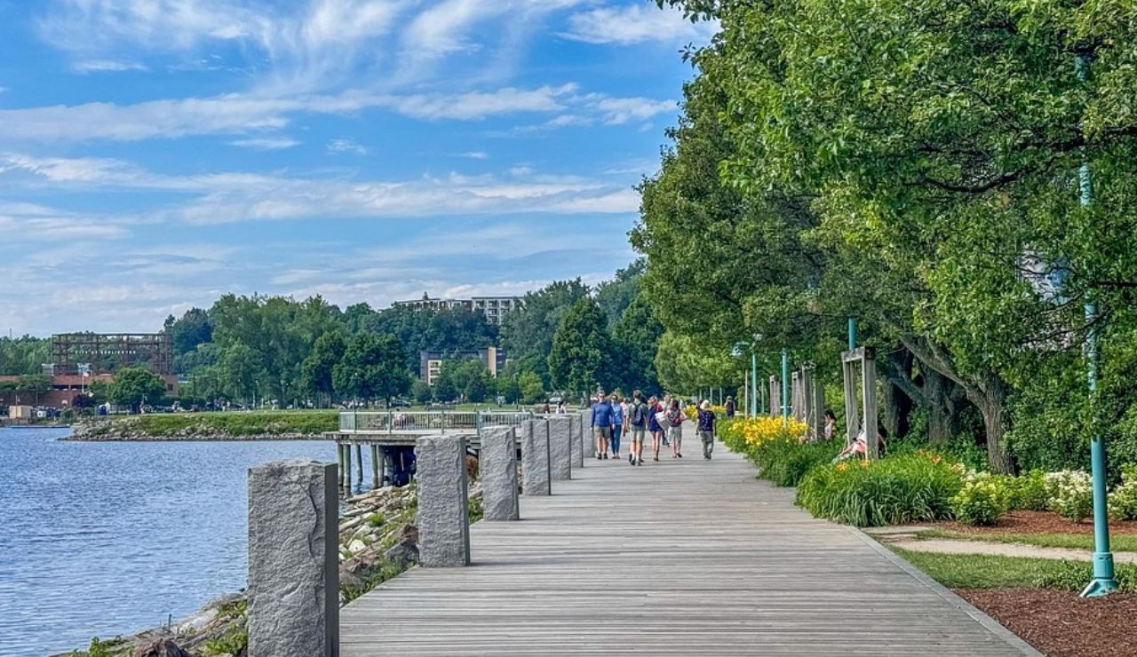 Scenic lakeside boardwalk with people walking, surrounded by greenery, flowers, and a view of buildings in the distance under a partly cloudy sky.