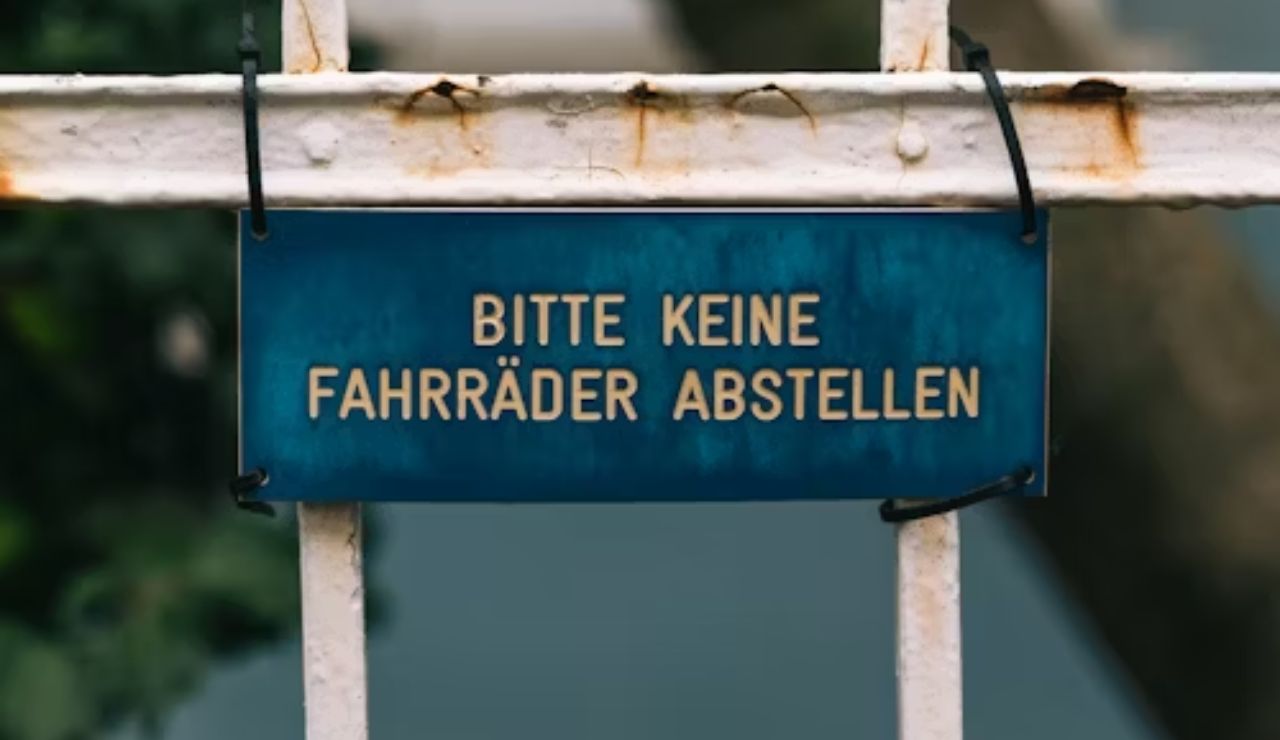 Blue metal sign on a white fence with German text "Bitte keine Fahrräder abstellen," meaning "Please do not park bicycles."