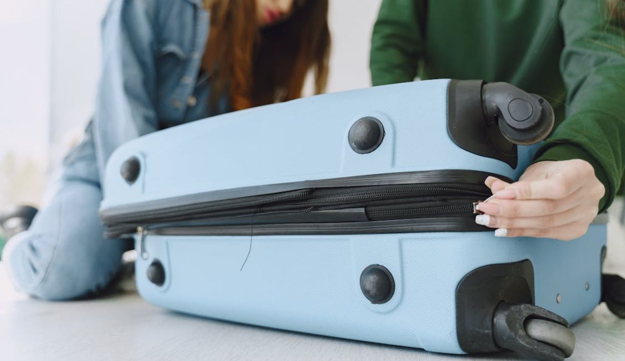 A person closing the zipper of a blue suitcase while another person is nearby.