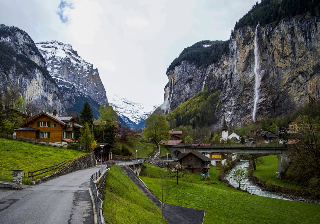 Scenic alpine village in Lauterbrunnen, Switzerland with wooden houses, snowy peaks, and tall waterfalls cascading down cliffs.