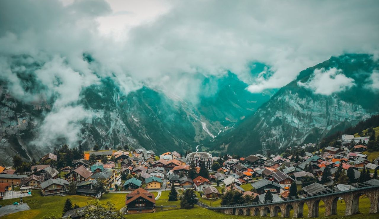A scenic mountain village nestled in a lush valley, surrounded by steep cliffs and misty clouds, with a curved stone viaduct in the foreground.