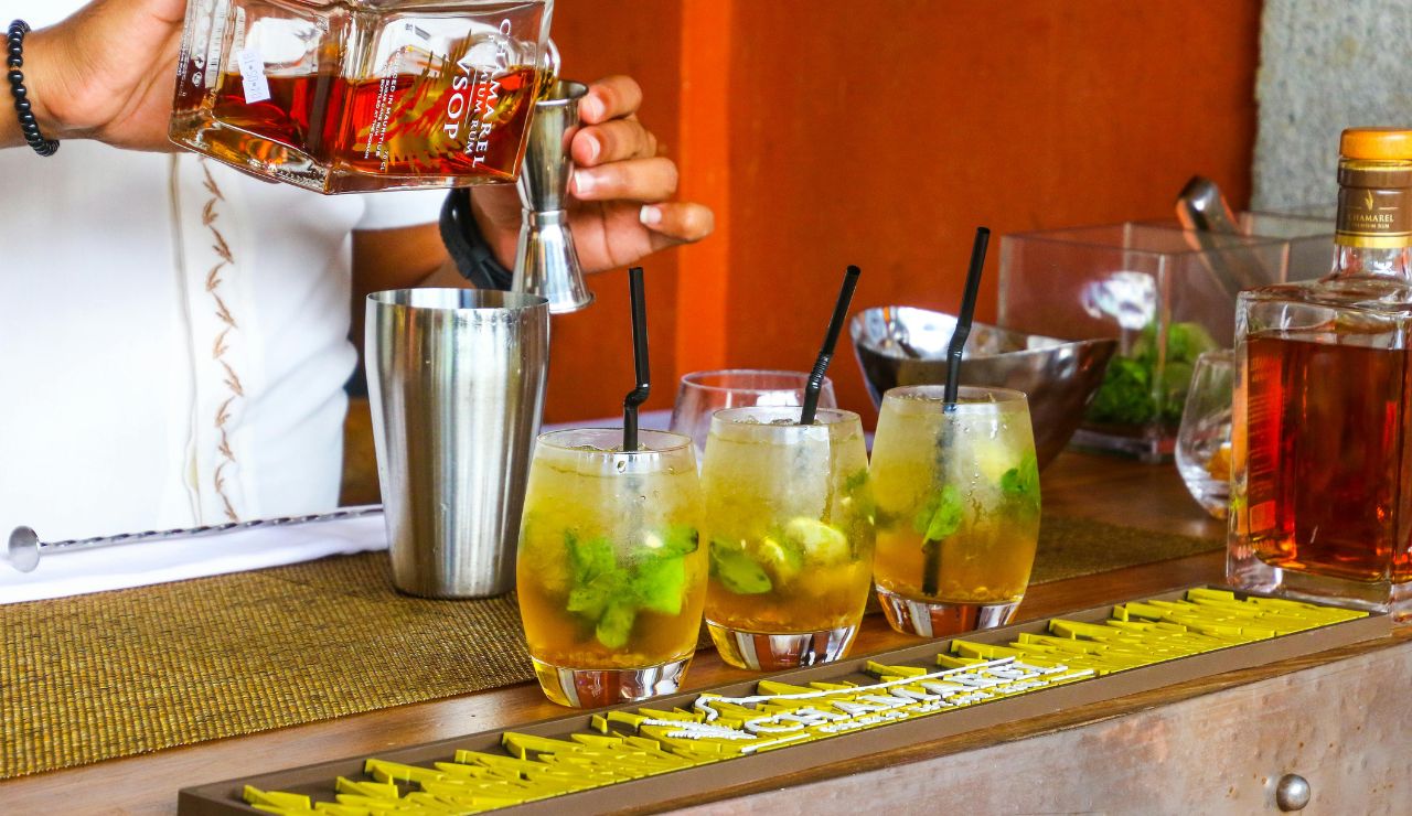 A bartender pours liquor into a jigger next to three mojitos on a bar counter, with cocktail tools and ingredients in the background.