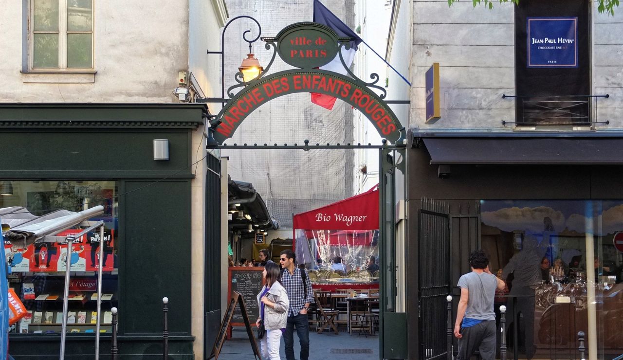 People walk beneath the Marché des Enfants Rouges sign in Paris, with shops and a restaurant visible inside.