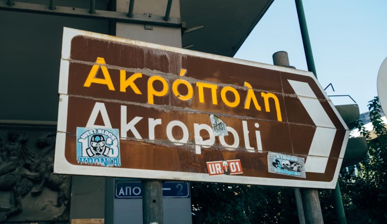 Weathered street sign in Greek and English pointing toward the Acropolis, with various stickers attached.