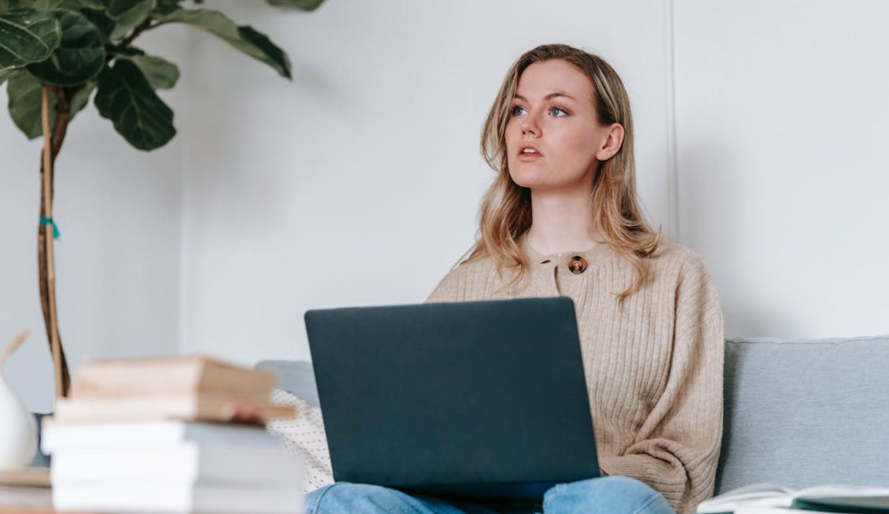 Woman sitting on a couch with a laptop on her lap, looking thoughtful, surrounded by books.