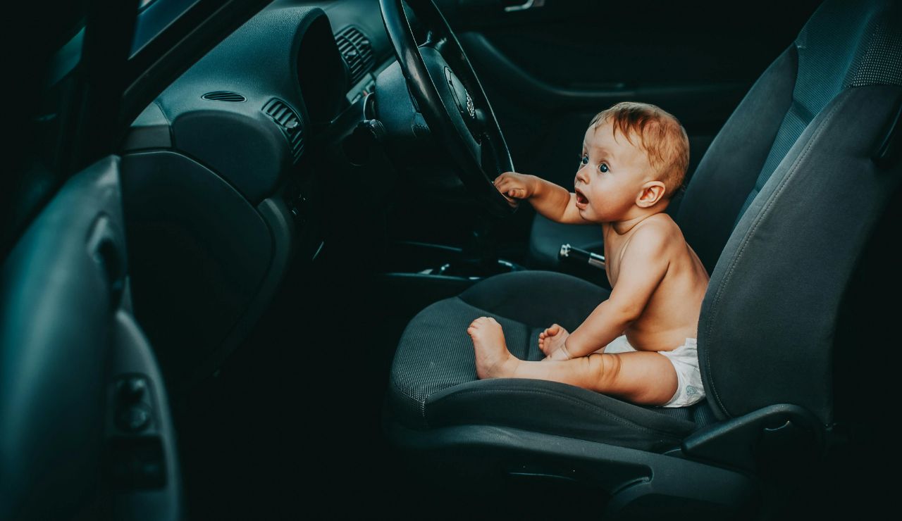 Baby in a diaper sitting in the driver's seat of a car, reaching toward the steering wheel with curiosity.