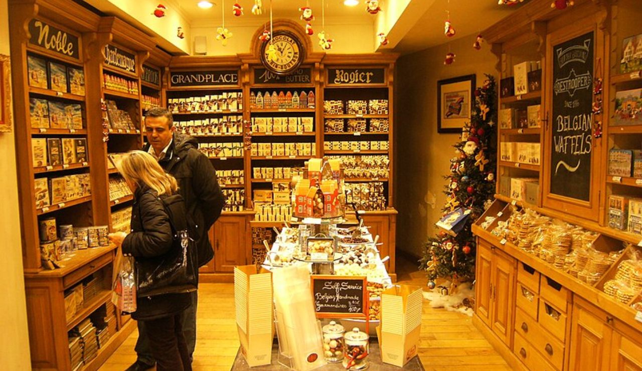 Interior of a cozy Belgian chocolate and waffle shop with wooden shelves, festive decorations, and a couple browsing the products.