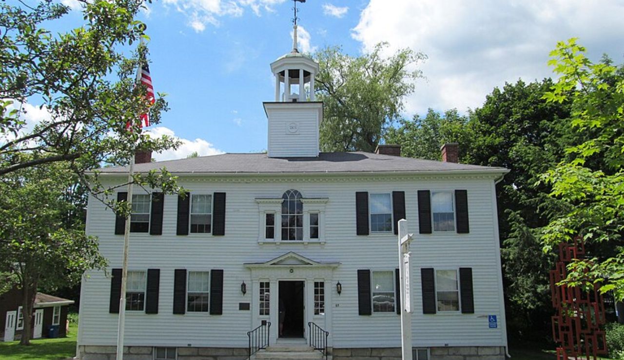 Historic white colonial-style building with black shutters, a central cupola, and an American flag out front.