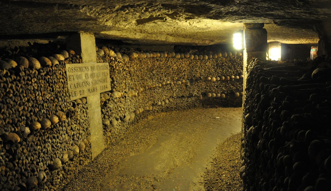 A dimly lit underground corridor lined with stacked human skulls and bones features a stone plaque mounted on the left wall.