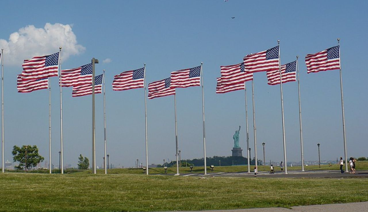 Row of American flags waving in the wind, with the Statue of Liberty visible in the distance under a clear blue sky.