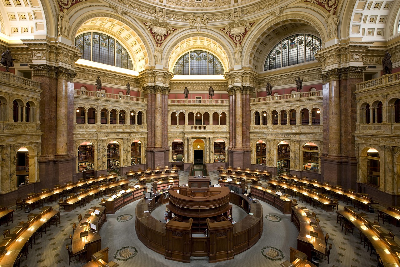 Grand interior view of a historic library reading room with arched windows, ornate columns, and rows of wooden desks arranged in a circular pattern around a central desk.