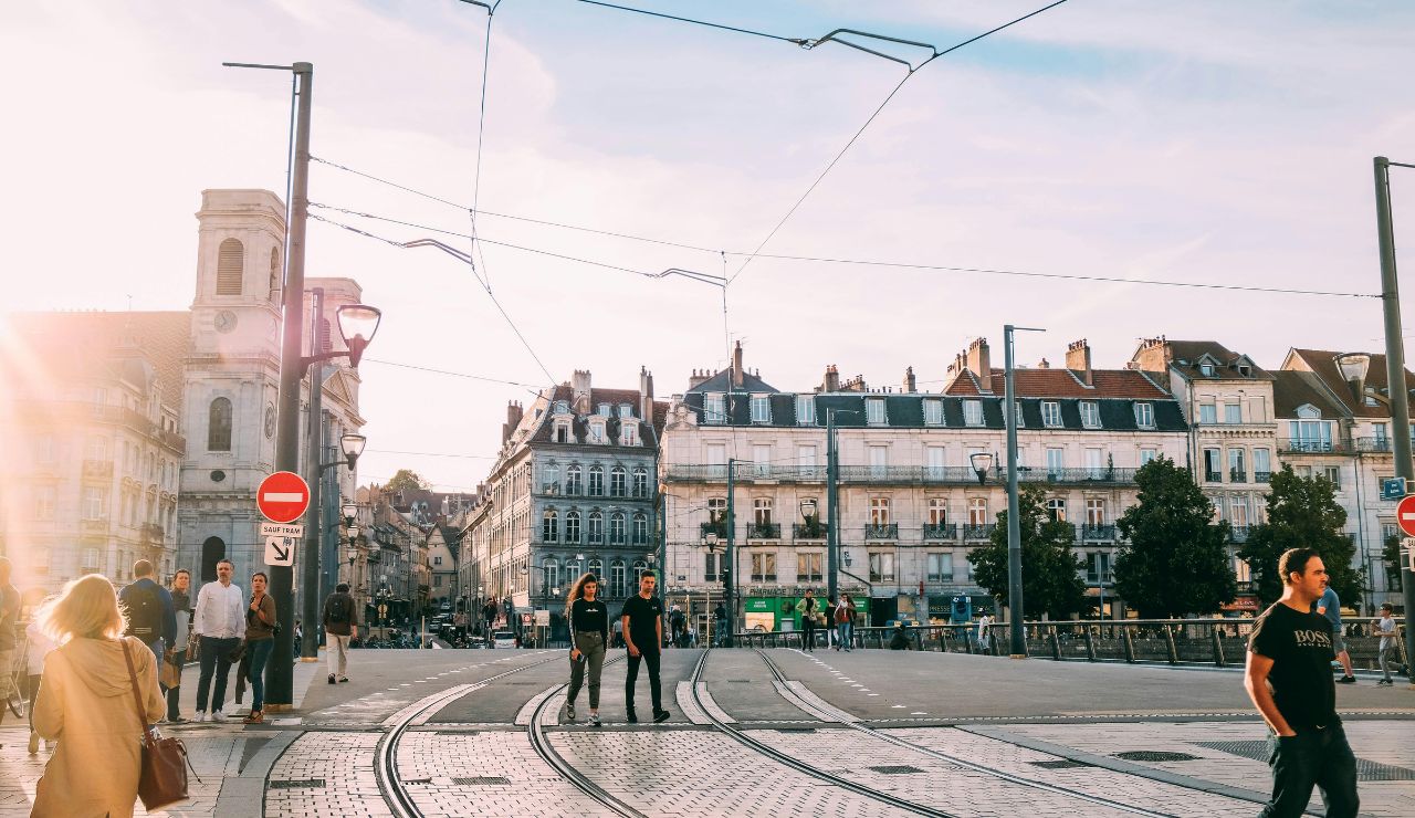 People walking along tram tracks in a sunlit European city square with historic buildings and overhead wires.
