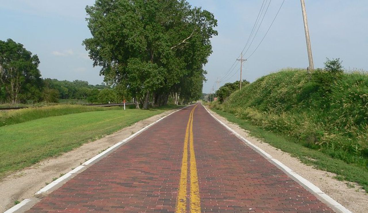 Historic brick-paved road lined with grass and trees, featuring double yellow center lines and utility poles along the side.