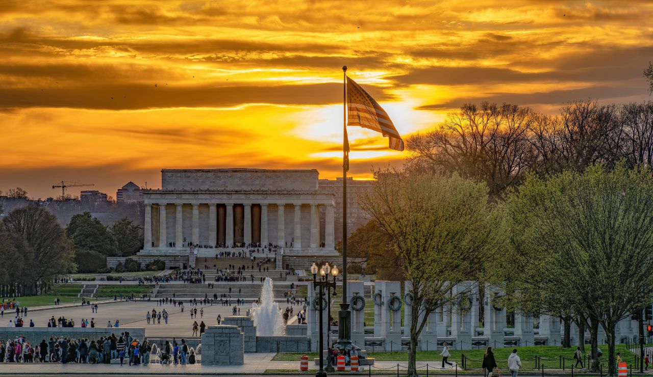 Sunset view of the Lincoln Memorial with a waving American flag in the foreground, surrounded by trees, fountains, and visitors in Washington, D.C.