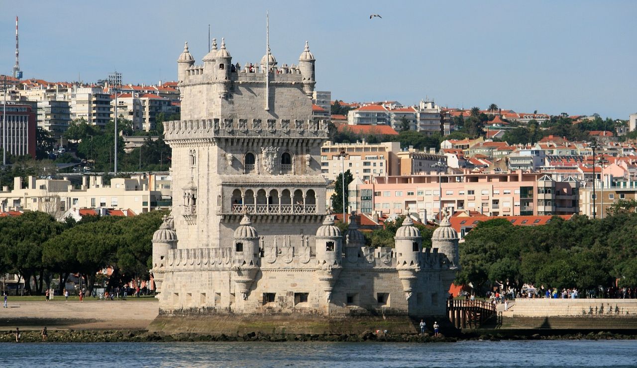 Historic stone tower on the waterfront with a backdrop of modern city buildings, surrounded by greenery and visitors.