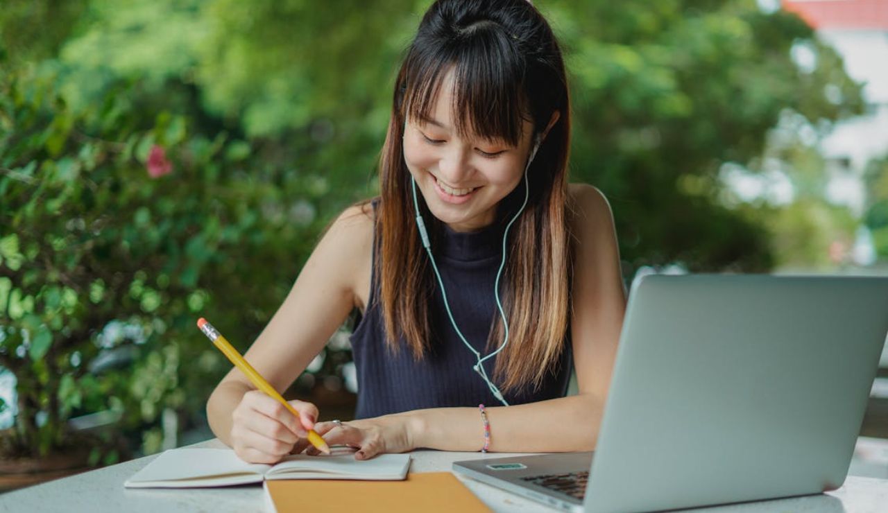 Smiling woman studying outdoors with a laptop, notebook, and earphones. Ask ChatGPT