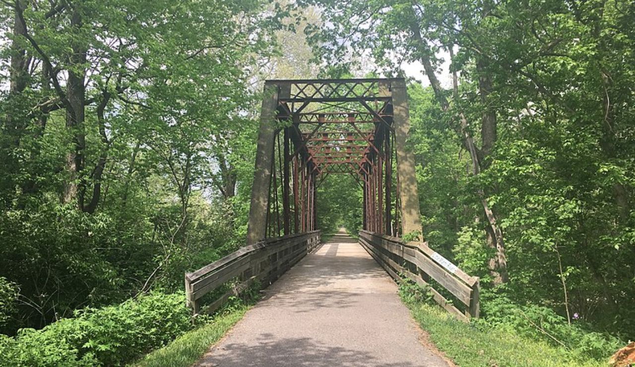 Paved bike and walking trail crossing a historic metal truss bridge surrounded by dense green forest on a sunny day.