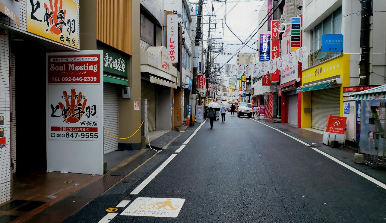 A wet Japanese city street with closed shops, colorful signs, and a few people walking under umbrellas.